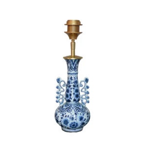 Productfoto van een Delftsblauwe lampenvoet zonder kap tegen een witte achtergrond. De vaasvormige voet is rijkelijk beschilderd met blauwe bloemen en bladmotieven, en aan weerszijden versierd met opvallende opengewerkte krullen. De messing fitting vormt een warme tegenhanger.