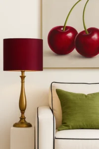Elegante tafellamp met kersenrode cilindervormige lampenkap van zijde op een goudkleurige voet, geplaatst naast een crèmekleurige fauteuil met groen sierkussen. Op de achtergrond hangt een schilderij van twee rode kersen tegen een neutrale muur.
