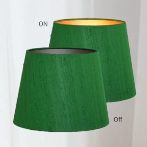 Twee identieke groene conische lampenkappen van wilde zijde naast elkaar. De linker is verlicht en laat een warme gloed zien dankzij de goudkleurige binnenzijde. De rechterkap is uitgeschakeld en toont de originele diepe groene kleur. Tekst bij de kappen geeft "ON" en "OFF" aan. Gefotografeerd tegen een lichte achtergrond.