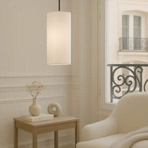 Interieurbeeld met een witte, smalle en hoge lampenkap boven een houten bijzettafel met kaars en vaas. De lamp hangt voor een raam met siersmeedwerk.