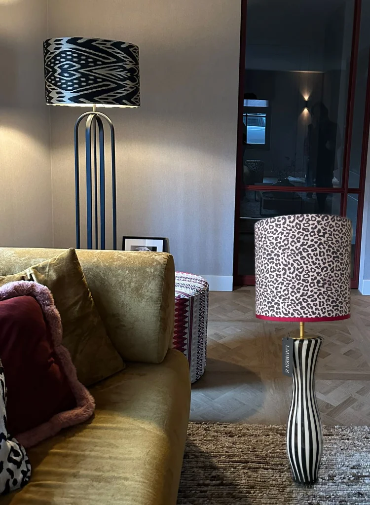 Interieurfoto van een moderne woonkamer met links een hoge zwarte metalen vloerlamp met een grote ronde lampenkap in zwart-wit ikatpatroon, en rechts op de voorgrond een tafellamp met een zwart-wit gestreepte voet en een opvallende luipaardprint kap met bordeauxrode afwerking. De combinatie van patronen en kleuren zorgt voor een eclectische en stijlvolle sfeer.