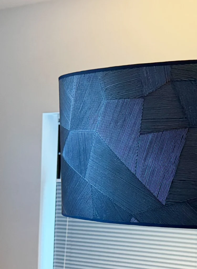 Detailfoto van een ronde lampenkap bekleed met diepblauw Arte-behang, waarin een geometrisch patroon van geweven lijnen zichtbaar is door het invallende daglicht. De structuur en textuur van het behang geven een luxe en modern effect.