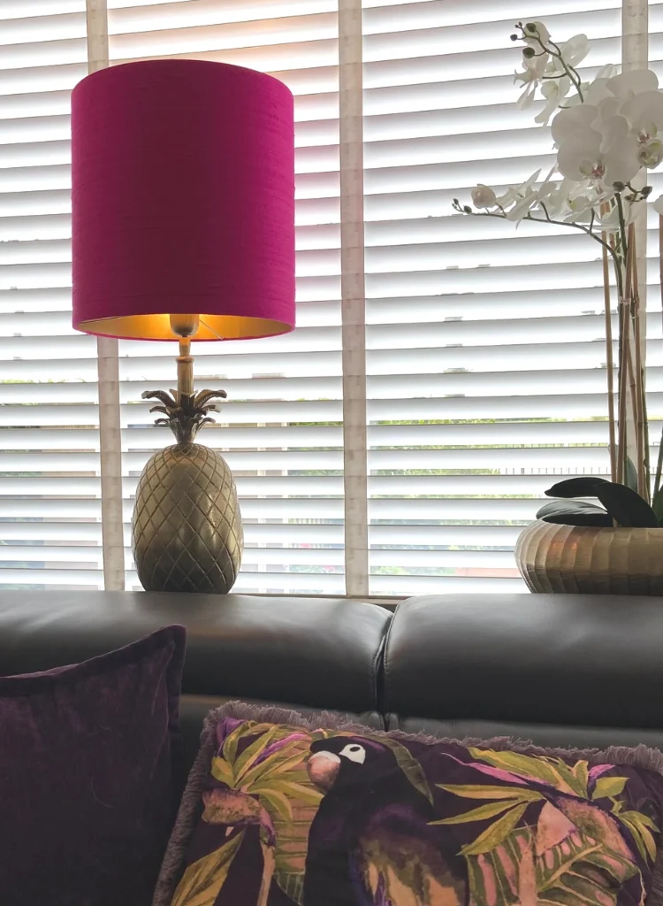 Een hoge, ronde lampenkap van fuchsiaroze zijde staat op een goudkleurige ananasvormige lampenvoet, geplaatst op een vensterbank in een restaurant. De achtergrond bestaat uit witte horizontale jaloezieën die zacht daglicht doorlaten. Rechts staat een grote vaas met witte orchideeën, links een donker paars kussen en een sierkussen met tropische print op een zwarte bank. De setting straalt luxe en speelsheid uit.