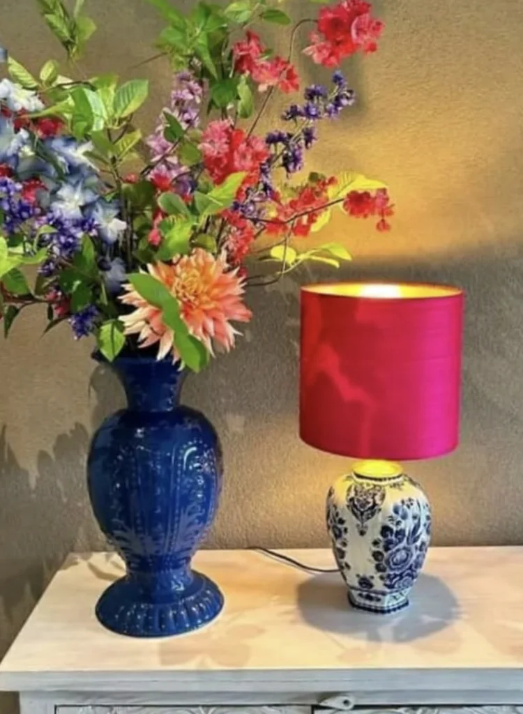 Een tafellamp met Delftsblauwe vaasvoet en felle cherryrode lampenkap staat op een lichte houten kast naast een grote blauwe vaas met kleurrijk zijden bloemenboeket.