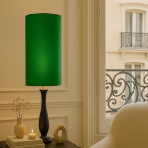 Hoge, cilindervormige lampenkap van handgeweven groene wilde zijde op een zwarte, elegante lampenvoet met goudkleurige fitting. De lamp staat op een houten bijzettafel in een klassiek Haussmann-interieur, naast keramieken objecten, een boek en een brandend kaarsje. De structuur van de zijde is zichtbaar en zorgt voor een luxe, warme gloed in het zachte daglicht.